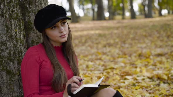 Pretty Thoughtful Girl Writing Down in a Notebook Near a Tree in Autumn Park
