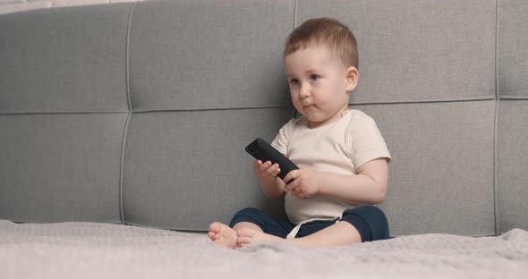 Baby Boy Sits on Bed and Holds Remote Control From the TV