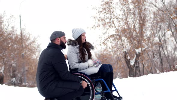 Happly Disabled Woman Sitting in Wheelchair is Talking to Beloved Husband at Park in Winter They are