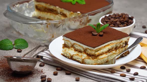 Traditional Italian Tiramisu dessert in glass baking dish and portion on grey concrete background