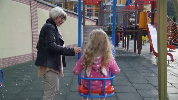 Back View Happy Carefree Granddaughter Sitting on Swings As Slim Beautiful Grandmother Swinging Kid