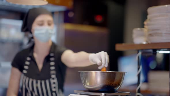 Metal Bowl at Front with Blurred Young Woman in Coronavirus Face Mask and Cook Hat Working at