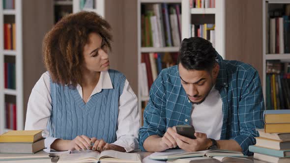 Two Diverse Students Sitting in Library Preparing for Exam Young Guy Enthusiastically Plays on Phone