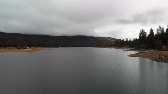 Aerial shot over smooth lake and pine trees, oncoming storm clouds cover mountains