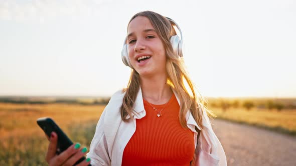 Young Happy Teenage Girl Walks Across Field Wearing Headphones and Sings Song