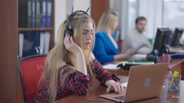 Portrait of Carefree Young Woman in Headphones Listening to Music Sitting in Office with Blurred