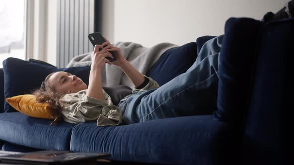 Young Smiling Woman Relaxing At Home Lying On Sofa Checking Social Media On Mobile Phone