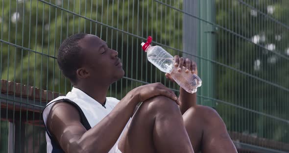 Young African American Streetball Player Taking Break at Outdoor Playground Sitting and Drinking