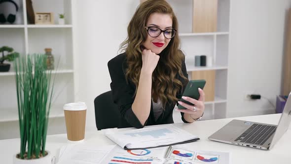 Business Lady in Glasses Dressed in Trendy Jacket with Brightly Red Lipstick Using Her Phone