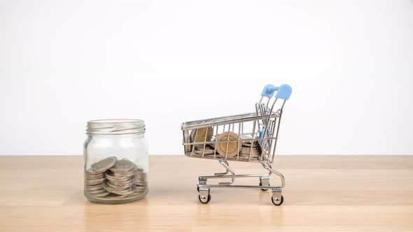 Stop motion animation shopping cart are carrying coins into a clear glass jar on wooden desk alt