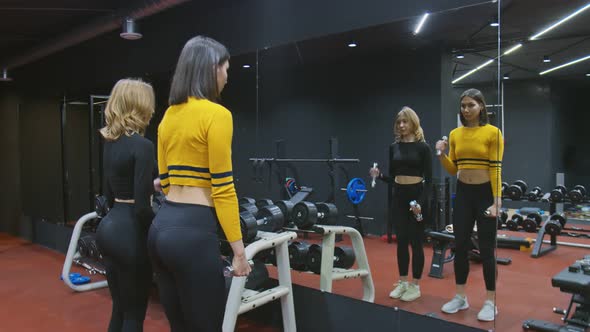 Two Beautiful Sexy Trainer Athletes Looks at Mirror and Doing Biceps Exercise with Two Dumbbells