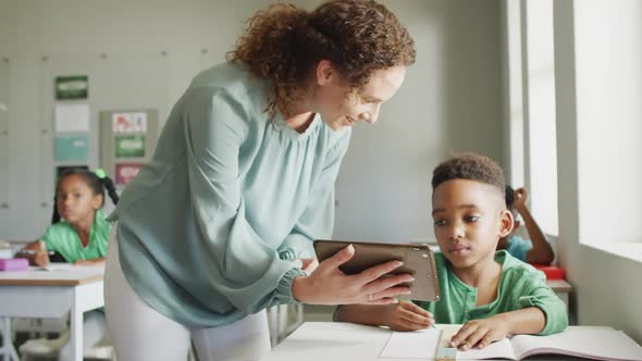 Video of happy caucasian female teacher explaining lesson on tablet to african american boy