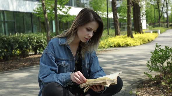 girl in denim jacket reading a book at alley near modern building of University