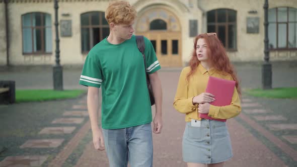 Positive Couple of Students Walking on Sunny University Yard and Chatting. Gogeous Redhead Woman and