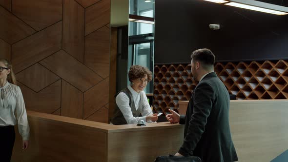 Male Guest Talking To Hotel Receptionist and Getting Key Card While Man and Woman Walking By