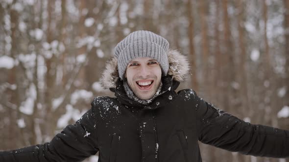 Positive Young Man in Winter Hat Breaks a Snowball with His Hands in the Forest