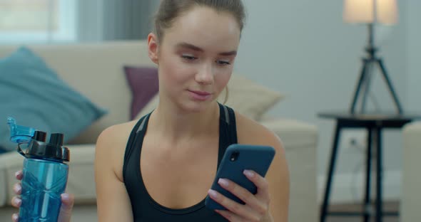 Sporty Woman Drinking Water and Use Phone While Resting After Training at Home. Healthy Lifestyle