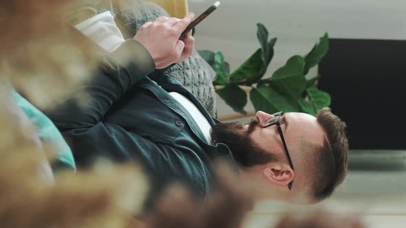 Millennial Bearded Man Sitting on Couch at Home Chatting Typing Message Using Cellular