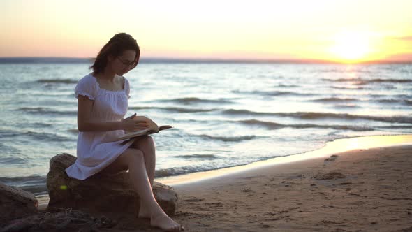 A Young Woman Sits on a Stone on the Beach By the Sea with a Book in Her Hands. A Girl in a White