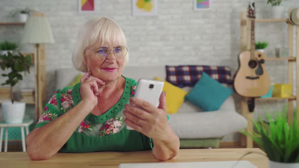 Thoughtful Elderly Woman Finds a Solution and Uses a Smartphone Sitting at the Table in the Living