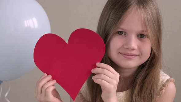 Close up portrait of cute little girl holding big red heart on background of air balloons