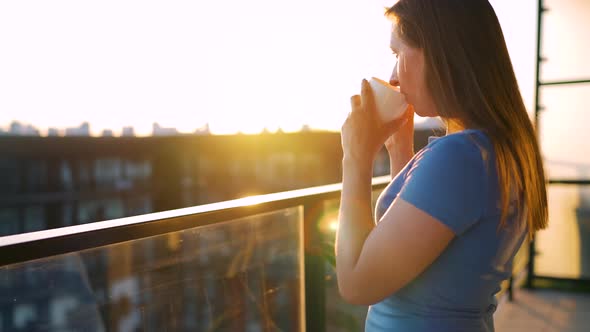 Woman with a Cup of Coffee Standing on the Balcony and Admire the Sunset