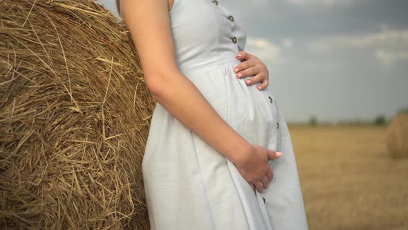 A Young Pregnant Woman Stands Near a Haystack in a Field