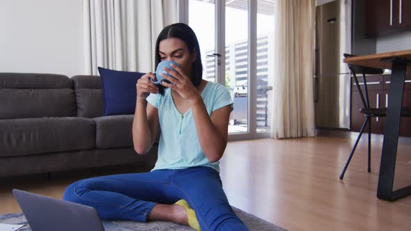 Mixed race gender fluid person sitting on floor drinking coffee and using laptop at home