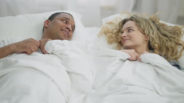 Young Happy Millennial Couple Hiding Under White Blanket Cuddling Lying in Bed