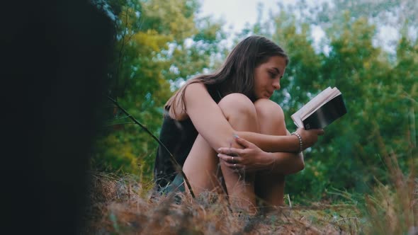 Young Woman Reads a Book Sitting on the Grass in Nature at Summer Day