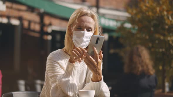 Mature Woman in Mask Enjoying Tasty Coffee Using Cellphone in Cafe on Sunny Day.