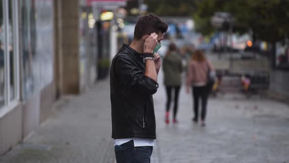 A young man in a leatherette jacket and jeans with a modern hairstyle on a busy street and puts on a