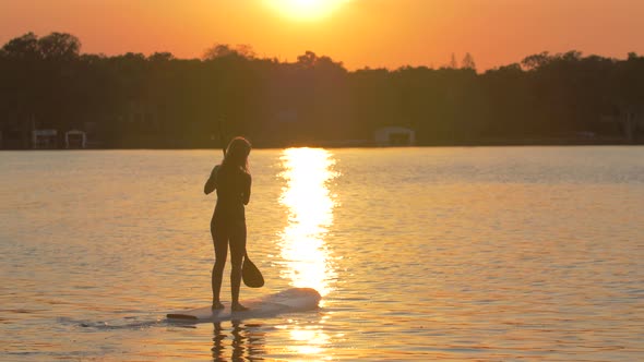 A young woman sup stand-up paddleboarding on a lake at sunset.