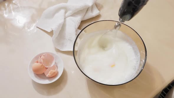 In a glass bowl on the table, the dough is whipped with a blender. Eggshells are in a small bowl