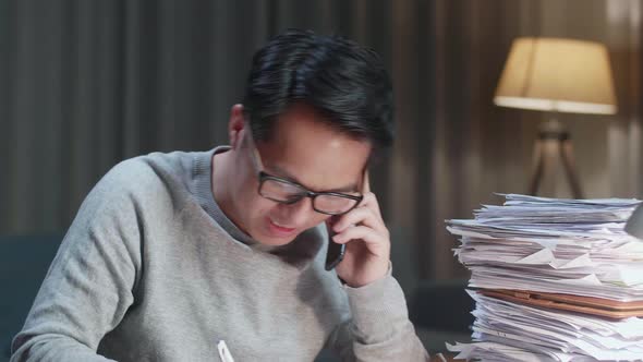 Close Up Of Asian Man Talking On Phone And Taking Note While Working Hard With Documents At Home
