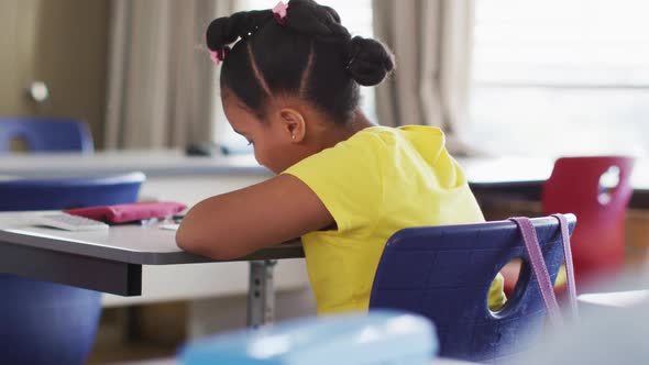 Portrait of happy mixed race schoolgirl sitting at classroom, making notes, looking at camera