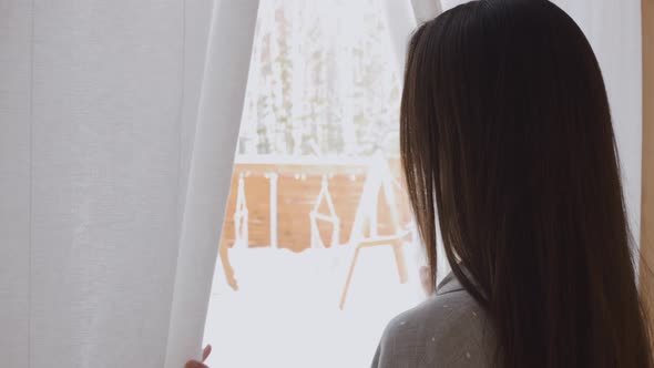 A Brunette Woman Opens the Curtains in the Morning and Looks at the Backyard Covered with Fresh Snow