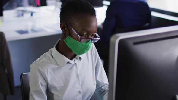 African american woman wearing face mask using computer while sitting on her desk at modern office