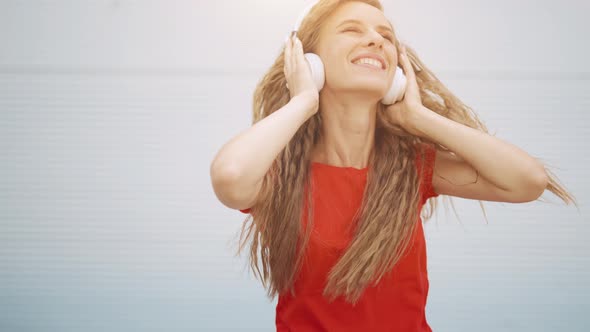A Young Girl in Headphones is Listening to Music and Amusingly Dancing on a Light Background