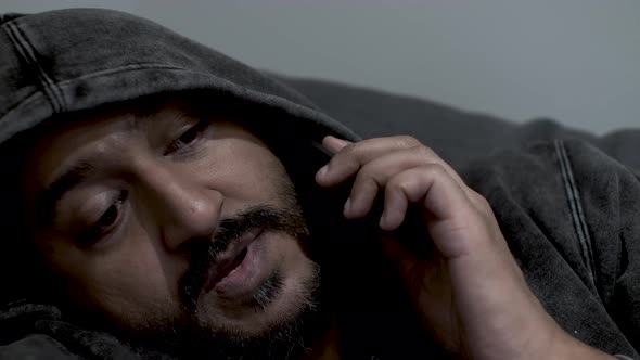 Close up shot of the face of an Indian man wearing a hoodie half asleep talking on his mobile phone,