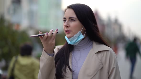 Pretty Young Woman with Protective Face Mask While Using Smart Phone and Sending Voice Message in