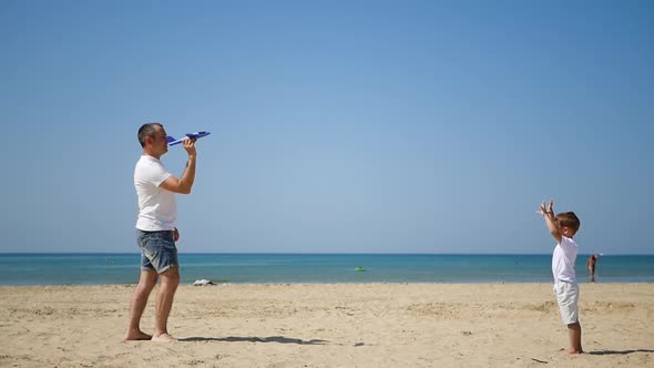A Happy Father and Son on the Sea Sandy Beach Are Facing Each Other and Launching a Toy Plane. The