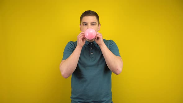 A Young Man Inflates a Pink Balloon with His Mouth on a Yellow Background. Man in a Green Polo.