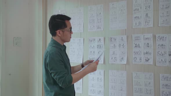 Young Creative Asian Designer Looks At The Storyboard Sketches Covering His Wall