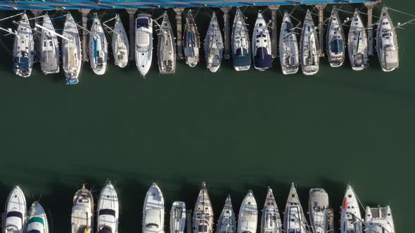 Several yachts and motorboats lie moored in crossed hawsers on several long blue jetties on the calm