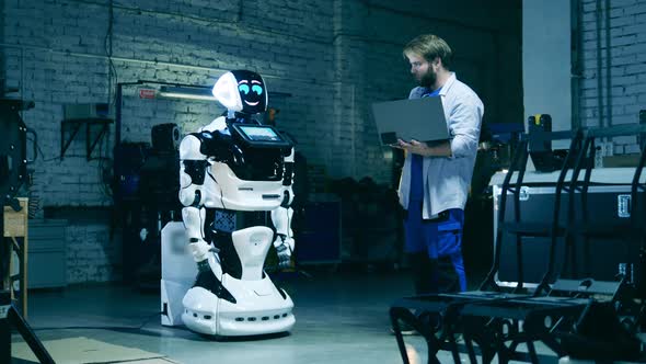 Serviceman with a Laptop is Observing a Modern Robot