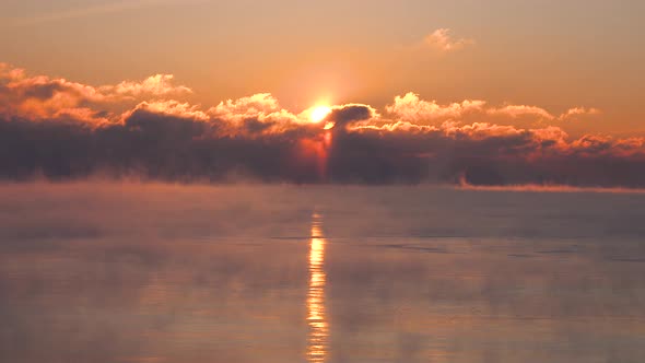 bright yellow red and orange sunrise through dark clouds over a very cold lake as steam rises in the