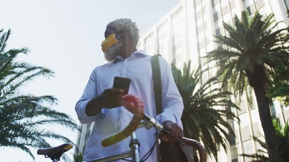 African american senior man wearing face mask with bicycle using smartphone and looking around in co