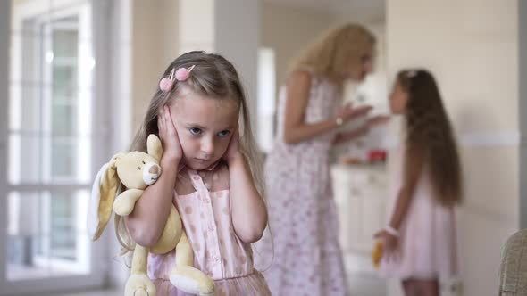 Pretty Little Caucasian Girl Covering Ears with Hands As Blurred Mother and Sister Arguing at