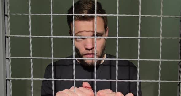 A Man In Handcuffs In The Prison Cell Stands Upset, Holding On To The Bars. Dangerous Criminal